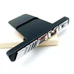 Puede incluir: Un emblema de plástico negro con un logotipo "AMG" en cromo plateado. El logotipo está en relieve y tiene un efecto tridimensional.