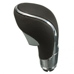 Op de afbeelding: Een donkerbruine auto versnellingspookknop met een chromen accent. De boven- en zijkanten zijn donkerbruin en de onderkant en een gebogen accent zijn chroom. De versnellingspook is ontworpen voor een auto.