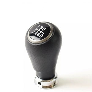 Op de afbeelding: Zwarte leren versnellingspookknop met een zilveren ring en een wit 5-versnellingschema met de letter "R" voor achteruit.