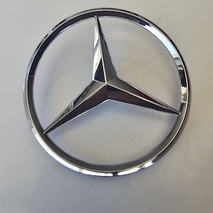 以下が含まれることがあります： クロムメッキのメルセデス・ベンツエンブレム。円形のフレーム内に三芒星が描かれています。エンブレムは反射性の金属仕上げで、白い背景に配置されています。星の先端はシャープで角張っており、モダンな美しさを生み出しています。