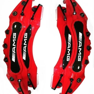 Könnte beinhalten: Zwei rote AMG-Bremssättel mit schwarzen Akzenten und dem weißen AMG-Logo. Die Bremssättel sind für den Automobilbereich konzipiert und vor weißem Hintergrund dargestellt. Sie haben mehrere Schraubenlöcher und schwarze Metallleitungen.