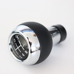 Op de afbeelding: Een zwarte lederen en chromen versnellingspookknop met een 5-versnellingschema, inclusief achteruit.