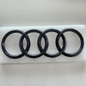 Puede incluir: Un emblema Audi negro con cuatro anillos entrelazados. El emblema está hecho de metal y tiene un acabado brillante.