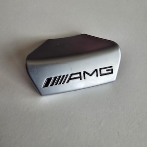 May include: A silver AMG emblem with black lettering and a distinctive shape. The emblem features the AMG logo and a stylized design. Likely a car accessory or part, it is presented on a neutral background.