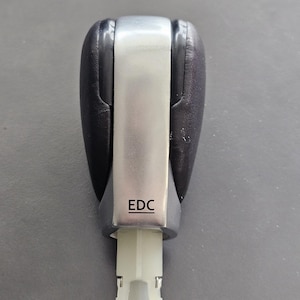 Peut inclure: Un pommeau de levier de vitesses noir et argent avec les lettres "EDC" imprimées sur le devant. Le pommeau de levier de vitesses a un centre argenté rectangulaire et des côtés noirs. La base est blanche.