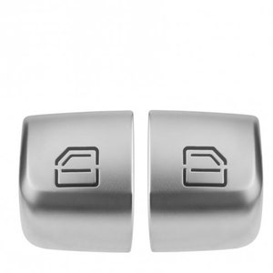 May include: Two silver car window switches with a door icon on each. The switches are rectangular and have a slightly rounded edge.