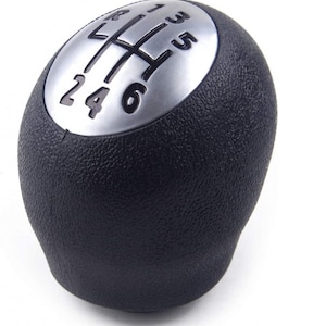 Peut inclure: Un pommeau de levier de vitesses noir avec un dessus argenté. Le dessus présente un motif de vitesses avec les chiffres 1, 2, 3, 4, 5 et 6.