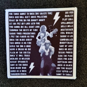 Puede incluir: Un parche bordado en blanco y negro con un borde blanco. El parche presenta una banda con dos miembros, uno sobre los hombros del otro, tocando guitarras. El parche tiene el texto "FOR THOSE ABOUT TO ROCK (WE SALUTE YOU)" y una lista de títulos de canciones de los álbumes de la banda.