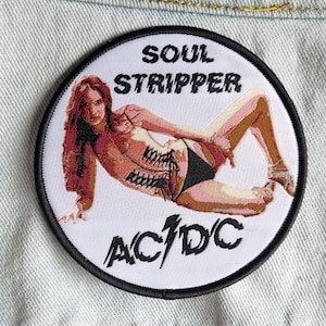 Peut inclure: Un écusson rond blanc avec une bordure noire représentant une femme en corset noir et blanc avec le texte "Soul Stripper AC/DC" en noir.
