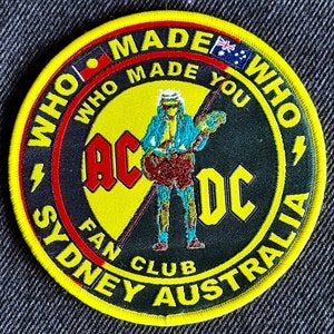 May include: A yellow and black round patch with the text "Who Made Who" and "AC/DC Fan Club Sydney Australia" around the edge. The centre of the patch features a cartoon image of a person playing a guitar with the text "AC/DC" in red and black.