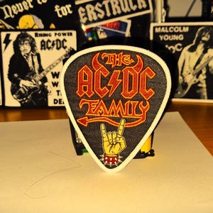 Peut inclure: Un &eacute;cusson brod&eacute; noir et rouge en forme de m&eacute;diator de guitare avec le texte "The AC/DC Family" et un &eacute;clair.
