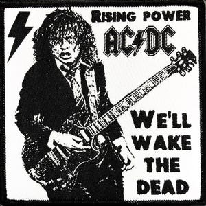 May include: Black and white patch featuring a graphic of a musician playing a guitar. The text "Rising Power AC/DC We'll Wake The Dead" is printed on the patch.