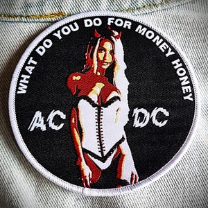 May include: A black and white embroidered patch with a red devil woman wearing a white corset. The text around the image reads "What do you do for money honey" and "AC/DC".