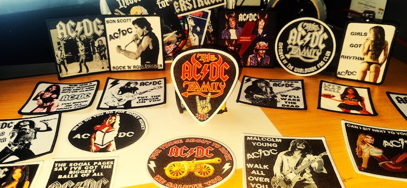 Peut inclure: Une collection de patchs et d'autocollants de groupe AC/DC color&eacute;s. Les patchs pr&eacute;sentent divers membres du groupe, des pochettes d'album et des paroles. Les autocollants sont principalement en noir et blanc avec quelques-uns en couleur. Les patchs et les autocollants sont tous sur une surface en bois.