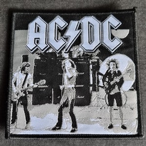 May include: Black and white embroidered patch featuring the band AC/DC with three band members in front of a microphone and speakers. The band's name is in white lettering with a lightning bolt.