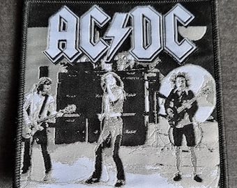 Op de afbeelding: Zwart-wit geborduurde patch met de band AC/DC met drie bandleden voor een microfoon en luidsprekers. De naam van de band staat in witte letters met een bliksem.