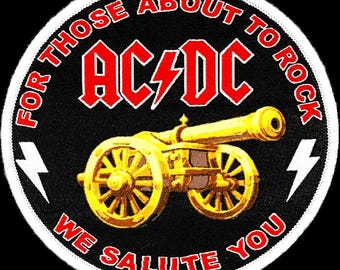 May include: A black and white circular patch with a white border. The centre of the patch features a gold cannon with a black barrel. The text "FOR THOSE ABOUT TO ROCK WE SALUTE YOU" is written in a white circle around the cannon. The text "AC/DC" is written in red letters above the cannon.