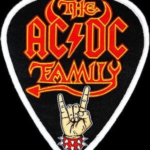 Puede incluir: Un parche negro en forma de púa de guitarra con un borde blanco. El parche presenta el texto "The AC/DC Family" en letras rojas y amarillas con una cola de diablo roja y un cuerno de diablo amarillo. Una mano de dibujos animados con una pulsera con púas está haciendo el gesto de rock and roll.