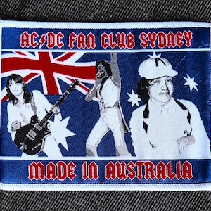 Op de afbeelding: Een blauwe en witte patch met de tekst "AC/DC Fan Club Sydney" en "Made in Australia". De patch toont een afbeelding van drie mannen, één speelt gitaar, één zingt en één draagt ​​een witte hoed.