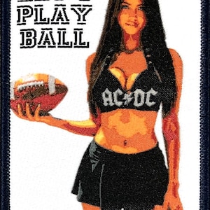 Puede incluir: Un parche blanco con borde azul que muestra a una mujer sosteniendo un balón de fútbol. La mujer lleva una falda negra, un top negro con el texto "AC/DC" en blanco y tiene el pelo largo y negro. El texto "LET'S PLAY BALL" está impreso en negro en la parte superior del parche.