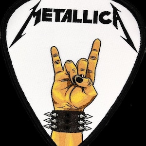 May include: White guitar pick shaped patch with black outline. The patch features the word "METALLICA" in black lettering and a yellow hand making the rock and roll hand gesture. The hand is wearing a black wristband with silver studs.