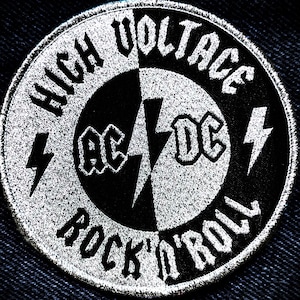 Puede incluir: Un parche circular blanco y negro con el texto "High Voltage" y "Rock 'n' Roll" alrededor del borde. El centro del parche presenta el logotipo de AC/DC con un rayo.