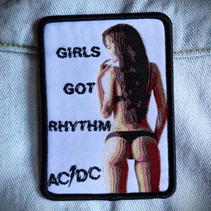 Puede incluir: Parche blanco con texto negro que dice "Girls Got Rhythm AC/DC". Una mujer con cabello negro largo está representada en la parte inferior de un bikini negro.