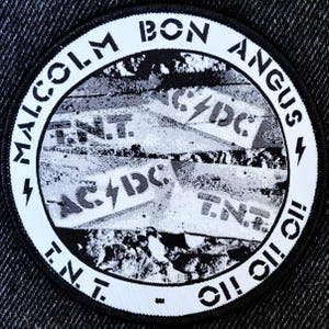 May include: A black and white circular patch with the text "Malcolm Bon Angus" around the edge. The centre of the patch features a collage of torn pieces of paper with the text "AC/DC" and "TNT" repeated multiple times.