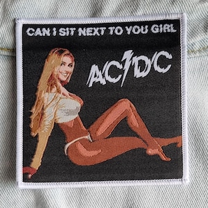 May include: A black and white embroidered patch with the text "CAN I SIT NEXT TO YOU GIRL" and the band name "AC/DC" in white. The patch features a woman in a white top and underwear, sitting with her legs crossed.