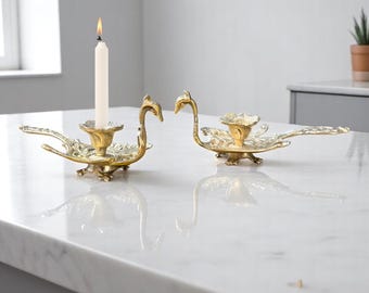 May include: Two antique brass swan candle holders. One has a lit white candle. The swan candle holders are on a white marble surface. The candle holders are in a room with a window and a potted plant.