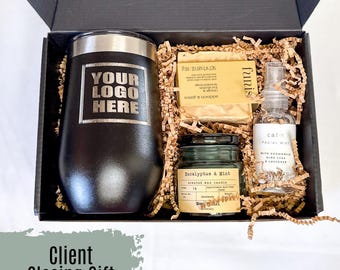 Pode incluir: Uma caixa de presente preta cheia de um copo preto e prateado, uma vela verde com um rótulo que diz "Eucalyptus & Mint", uma barra de sabonete com a palavra "funis" nela e um frasco de névoa facial com o rótulo "calin". O copo tem o texto "YOUR LOGO HERE" gravado.