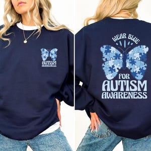 Pu&ograve; includere: Una felpa blu navy con una farfalla blu fatta di pezzi di puzzle sul retro. La farfalla ha il testo "Wear Blue For Autism Awareness". La parte anteriore della felpa ha una farfalla blu pi&ugrave; piccola con il testo "Autism Awareness".