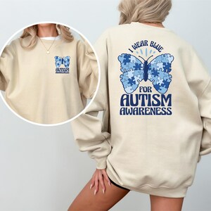 Pu&ograve; includere: Una felpa beige con una farfalla blu fatta di pezzi di puzzle sul retro. La farfalla ha il testo "Indossare il blu per la consapevolezza dell'autismo". La parte anteriore della felpa ha una farfalla blu pi&ugrave; piccola con il testo "Consapevolezza dell'autismo".