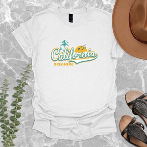 May include: White t-shirt with a graphic design of a palm tree and a sun with the text "California Dreaming" in a retro font.