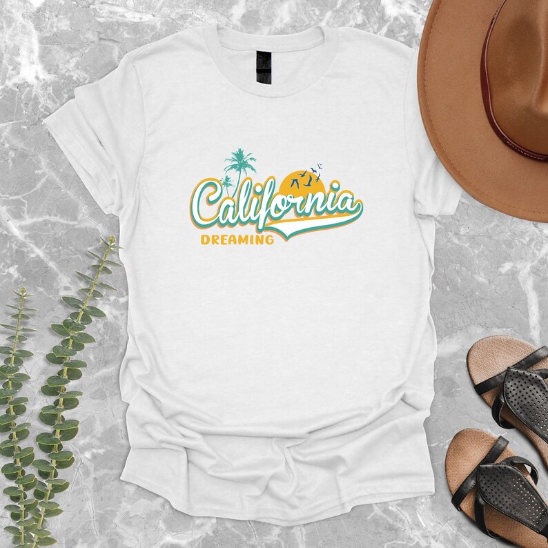 May include: White t-shirt with a graphic design of a palm tree and a sun with the text "California Dreaming" in a retro font.