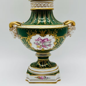 May include: A green and white porcelain urn with a matching lid, featuring gold trim, ram head handles, and a central cartouche depicting a pink ribbon, a book, and floral motifs. The base is decorated with a small floral spray and gold accents.