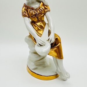 Könnte beinhalten: Porzellanfigur einer sitzenden Frau, die ein Saiteninstrument spielt. Die Figur trägt ein goldfarbenes Kleid und eine Kopfbedeckung. Sockel und Instrument sind weiß mit goldenen Akzenten.