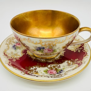 Può includere: Un set vintage di tazza e piattino da tè. La tazza ha un interno dorato e un esterno bianco con accenti floreali e dorati. Il piattino è bordeaux con bordi dorati e dettagli floreali.