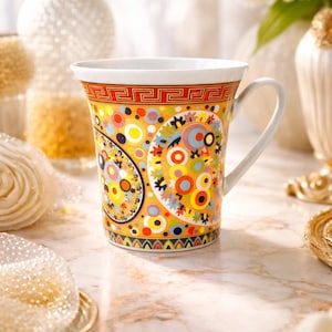 Könnte beinhalten: Eine dekorative Keramiktasse mit weißem Henkel und Rand. Die Tasse hat ein lebendiges, farbenfrohes Paisley-Design mit orangefarbenen, gelben, blauen und roten Akzenten. Die Tasse steht auf einer Marmoroberfläche, mit dekorativen Gegenständen im Hintergrund.