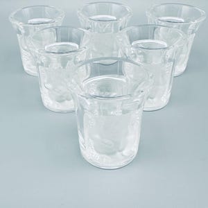 Könnte beinhalten: Sieben klare Schnapsgläser. Jedes Glas hat einen ausgestellten Rand und ein geätztes Design. Die Gläser sind kreisförmig auf einem hellblauen Hintergrund angeordnet. Ideal für Spirituosen.