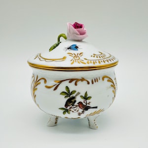 May include: A white porcelain lidded bowl with three feet, featuring gold filigree accents, a hand-painted scene of two birds on a branch, a small blue insect, and a sculpted pink rose finial on the lid with a green leaf detail.