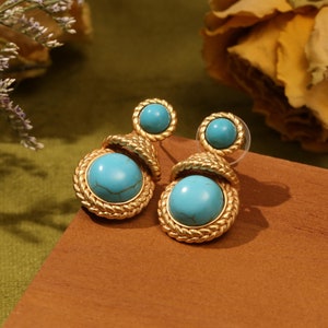 Può includere: Un paio di orecchini pendenti dorati con pietre blu turchese. Gli orecchini hanno un design intrecciato attorno alle pietre.