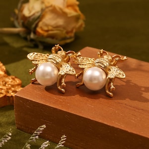 Peut inclure: Une paire de boucles d'oreilles en forme d'abeille dorées avec un accent de perle. Les abeilles sont détaillées avec des ailes texturées et un corps en relief.