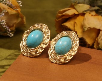 May include: A pair of gold-toned oval stud earrings with a turquoise stone center. The earrings have a textured, braided design around the stone. The earrings are displayed on a wooden surface.