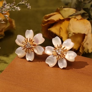 Puede incluir: Un par de pendientes de flores blancas con detalles dorados y brillantes transparentes en el centro.