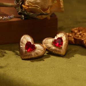 Könnte beinhalten: Ein Paar goldene herzförmige Ohrringe mit roten Edelstein-Akzenten.