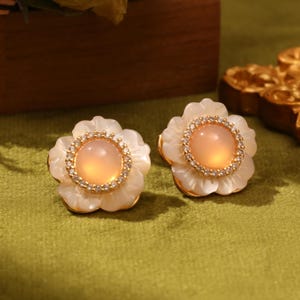 Könnte beinhalten: Ein Paar goldene Ohrringe in Blumenform mit einem rosafarbenen Stein in der Mitte und klaren Kristall-Akzenten.