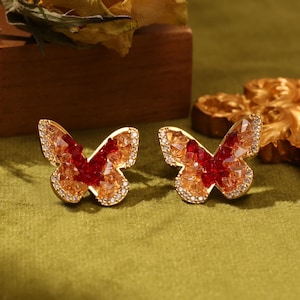 May include: A pair of gold butterfly earrings with red and clear crystal accents.