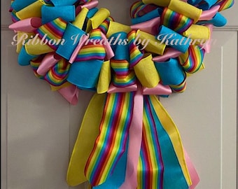 May include: A rainbow-colored wreath made with yellow, pink, and blue ribbon. The wreath has a bow with a rainbow stripe ribbon hanging down.