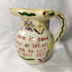 May include: A cream-coloured ceramic pitcher featuring a hand-painted green wavy line along the rim and handle. The body displays purple flower motifs, yellow accents, and brown text reading: there is good liquor, judge, or vicar, and not to spill any of your skill.